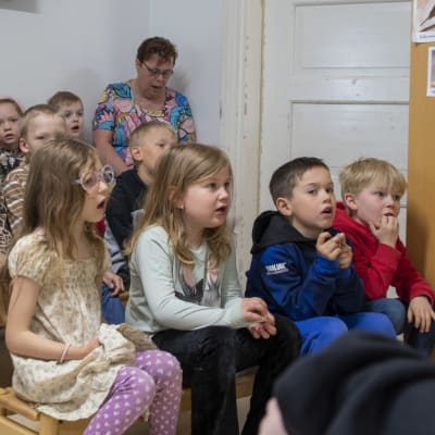 Förskolebarn sitter på bänkar i ett rum och sjunger tillsammans med en vuxen.