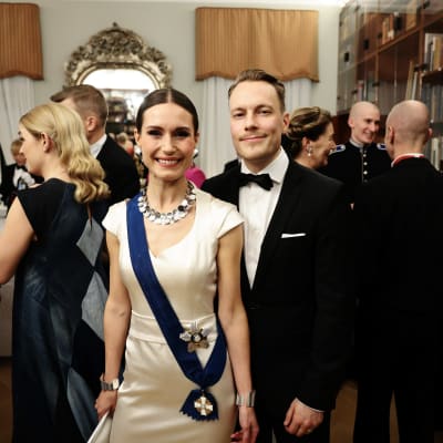 Sanna Marin i en vit klänning, tillsammans med hennes make Markus Räikkönen, i svart kostym.