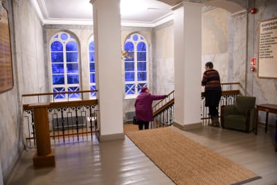 Två kvinnor i en stor aula med en trappa. Den ena kvinnan står på övre våningen, den andra är i trappan gående mot nedre våningen. De har ansiktena vända mot varandra och pratar. På motsatt vägg finns tre stora valvbågade fönster. Skymningen lyser blå genom fönstren.