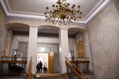 Vy från en trappa upp mot en stor aula. En guldfärgad takkrona hänger ner. Genom en öppen dörr syns tre personer i ett rum.