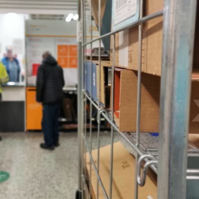 I förgrunden postpaket som står på en hylla. I bakgrunden personer som hämtar ut paket från en postkassa.