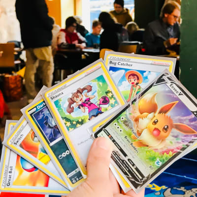Seitsemän erilaista Pokemon korttia henkilön kädessä. Taustalla näkyy muita Pokemon pelaajia.