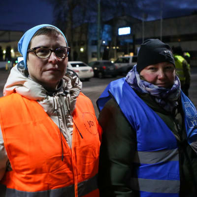 Två strejkvakter, två damer i orange och i blå reflexväst står i morgonmörkret utanför en fabrik. I bakgrunden syns fler strejkvakter i gula reflexvästar.