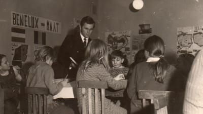 Nuori miesopettaja luokassa pulpeteissaan ahertavien pikkulasten keskellä, vanha valokuva.