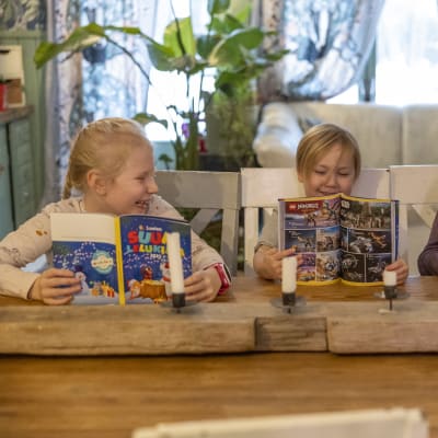 Kolme lasta selaa Lasten suurta lelukirjaa pöydän ääressä ja hymyilee tai nauraa.