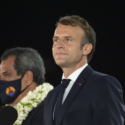 Bild på Frankrikes president Emmanuel Macron. Han står bredvid en person som applåderar, och en man i munskydd.