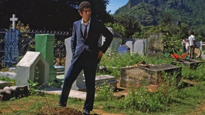 Mies (Näyttelijä Elliott Gould) seisoo hämmentyneen näköisenä hautausmaalla vasta umpeen luodun haudan ääressä. Kuva elokuvasta Pitkät jäähyväiset.