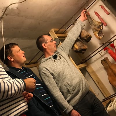 Matias Jungar, Michael Björklund och Pontus Berglund står nere i en källare och betraktar olika köttprodukter som hänger i band vid en vägg.