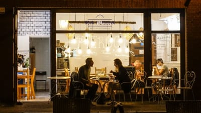 En restaurang fotograferad genom ett fönster. Två par syns äta mat vid separata bord.