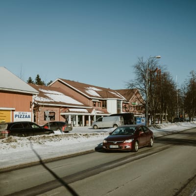 Rakennuksia, joiden vierestä kulkee tie. Tiellä ajaa auto. Useita autoja on taustalla parkissa. 