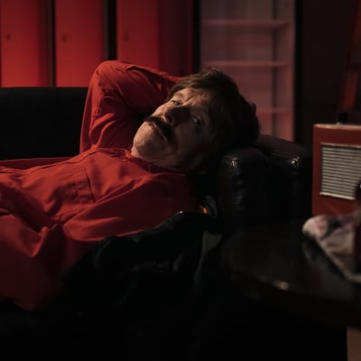 Viiksekäs mies (näyttelijä Juha Svahn) punaisessa kauluspaidassa makaa keinonahkasohvalla; kuva on mukaeltu Wim Wendersin elokuvan Paris, Texas kohtauksesta.