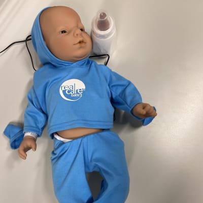En baby-docka iklädd en blå tröja och blåa byxor. En sladd löper från dockan ur bilden. Bredvid dockan en nappflaska.