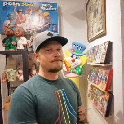 En man med keps och blond skäggväxt står framför en vägg fylld med spel och leksaker. På väggen finns ett bordshockeyspel tillverkat av företaget Bock-Plast.