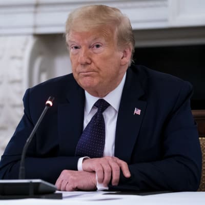 President Donald Trump sitter vid ett bord och håller armarna i kors. Han ser bekymrad ut. I bakgrunden syns USA:s flagga.