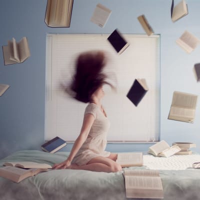 Nuori nainen istuu sängyllä, kirjoja lentelee hänen ympärillään.