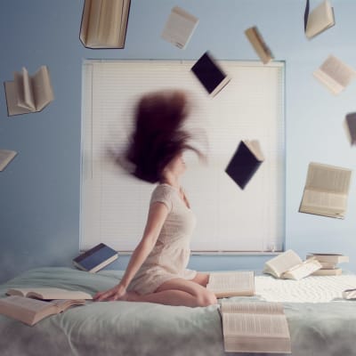 En kvinna på en säng med en drös böcker uppkastade i luften. Symbol för stress.