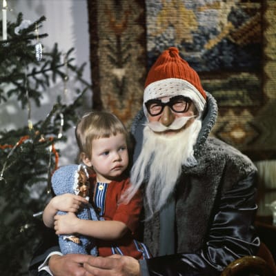 En liten flicka i julgubbens famn på julen. I bakgrunden en julgran.