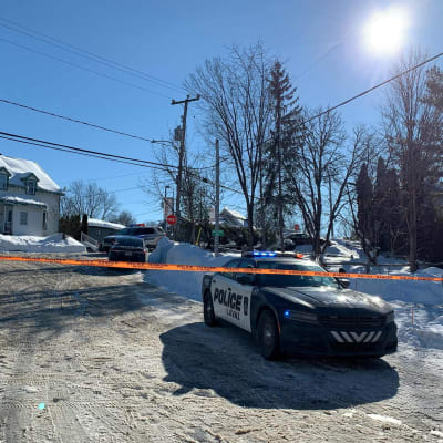 En polisbil står parkerad bakom ett orange avspärrningsband vid en snöig gata. En bit längre bort står flera bilar och ett ljust tvåvåningshus.