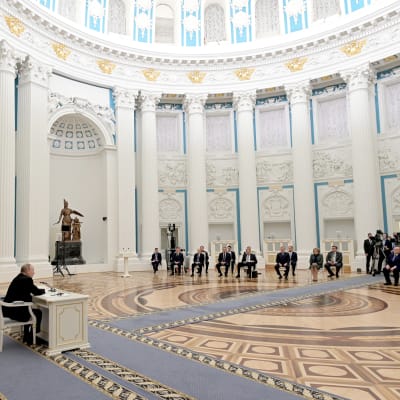 Rysslands president Vladimir Putin sitter i väldig sal. Putins närmaste rådgivare är samlade i en halvcirkel i salens andra ända.