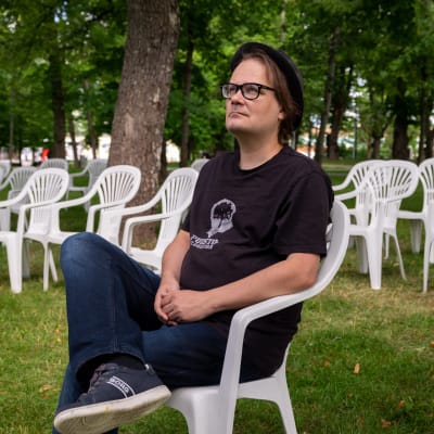 Mustaan t-paitaan ja farkkuihin pukeutunut mies istuu puistossa valkoisella muovipenkillä ja katsoo yläilmoihin. Miehellä tummasankaiset silmälasit ja hattu, taustalla vehreää puistoa.