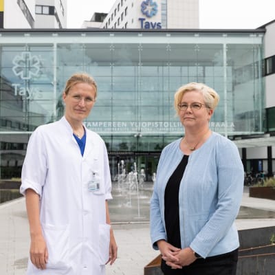 Kaksi naista seisoo Tampereen yliopistollisen sairaalan etupihalla. Toisella naisista on vaaleansininen neuletakki, toisella lääkärin asu. Taustalla näkyy pronssipatsas ja pääovet.