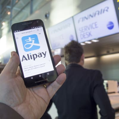 Alipays logotyp på en telefon framför Finnairs incheckningsdesk.