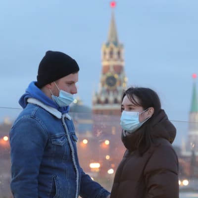 En man och en kvinna med munskydd. Mannen har dragit ner sitt skydd så att näsan inte är täckt. Personerna står framför Vasilijkatedralen som är ett känt landmärke i Moskva.