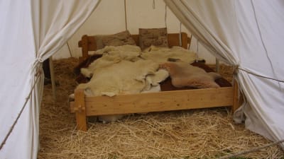 Träsäng fylld med djurfällar. Sängen står i ett tält på underlag av halm.