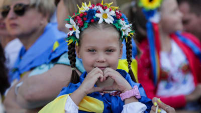 Liten flicka iklädd blomsterkrans invirad i Ukrainas flagga. Flickan ler och gör en hjärtsymbol med händerna.
