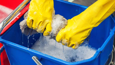 Två händer i gula gummihandskar vrider vatten ur en luddig golvmopp ner i en blå hink.