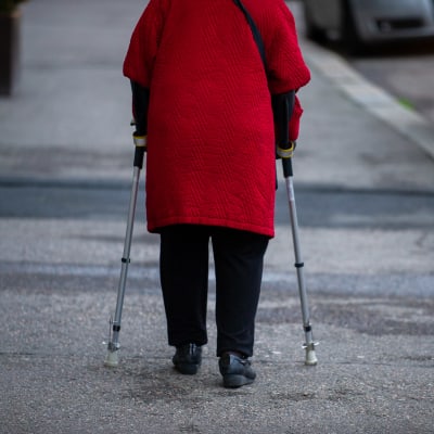 Äldre dam i röd kappa går gatan fram med hjälp av kryckor.