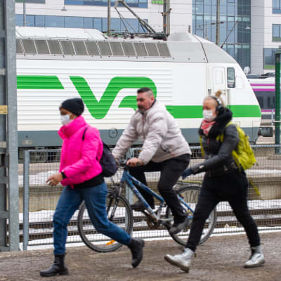 Två kvinnor i munskydd promenerar och en man utan munskydd på cykel på Helsingfors järnvägsstation 25.3.2021
