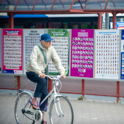 Man med skärmmössa cyklar förbi valplakat i Helsingfors.