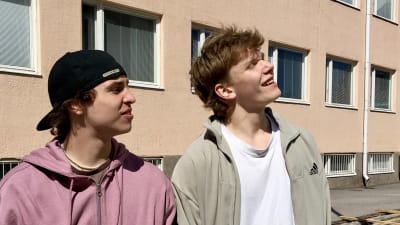 Två tonåriga pojkar på skolgård, den ena med lila tröja och keps, den andra med beige träningsjacka.