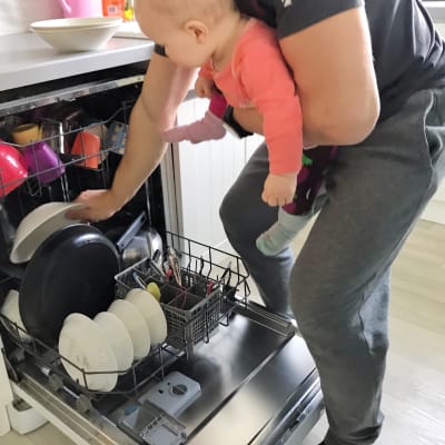 En pappa med sin baby i bärsele fyller diskmaskinen.