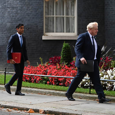 Tre män i kostym går efter varandra på en trottoar med en blomsterplantering och en byggnad i bakgrunden. Först går Boris Johsons, sedan följer Rishi Sunak och längst till vänster syns Sajid Javid.