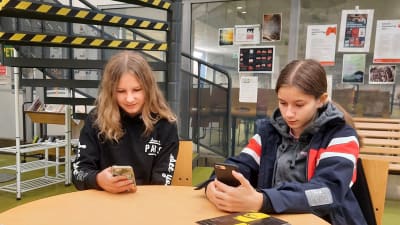 Två flickor sitter vid ett runt bord med mobiltelefonerna i handen. 