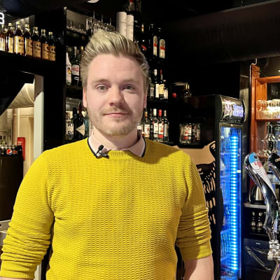 Baaritiskin takana seisova Väinö Kähkönen katsoo kohti kameraa. Taustalla näkyy hyllyssä erilaisia alkoholijuomia.