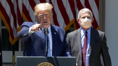 Donald Trump står vid ett talarpodium och håller presskonferens. I bakgrunden syns smittskyddsexperten Anthony Fauci som bär ansiktsskydd. Trump är utan.