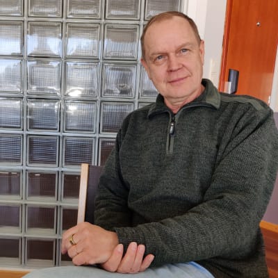 En man sitter på en stol med händerna i kors i knät. Han heter Markku Nikola.