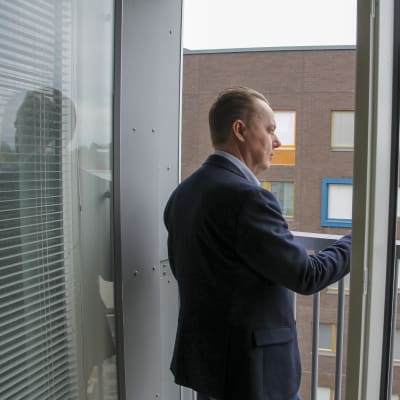 Siniseen pukuun pukeutunut mies seisoo parvekkeella ja katselee oikealle.