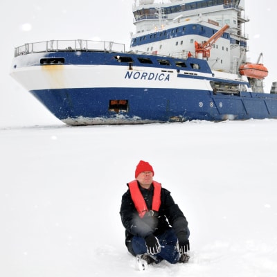 Ari Turunen istuu meren jäällä, taustalla jäänmurtaja Nordica.