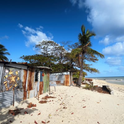 På bilden syns strandhytter, palmer och blå himmel på ön Masig som ligger i Torressundet norr om Australien.