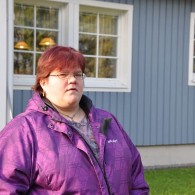 kvinna med kort rött hår och glasögon står utanförö sitt blåa egnahemshus.