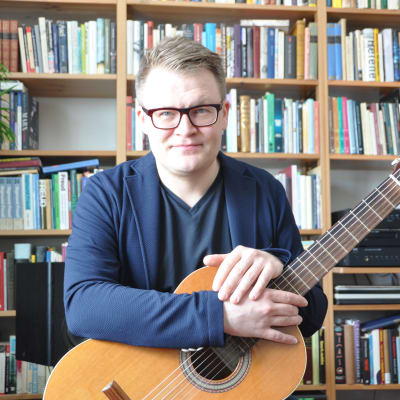Forskaren Juha Torvinen sitter i sitt vardagsrum med gitarren i knät