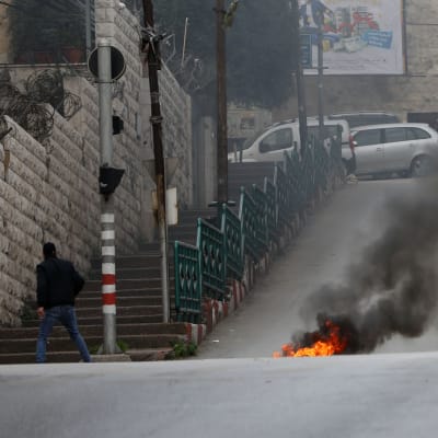 Ett militärfordon syns i bakgrunden av en stadsmiljö. I förgrunden brinner något på marken. En man rör sig bort från fordonet och elden medan han ser sig om över axeln.