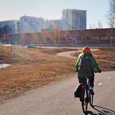 Vihreätakkinen pyöräilijä ajaa pyörätietä poispäin, taustalla pitkä punainen varikkotalo ja sen takana kerrostaloja.