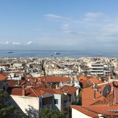 Vy över takåsarna i Thessaloniki där det syns tegelpannor på ljusa hus och på redden ligger några lastfartyg.