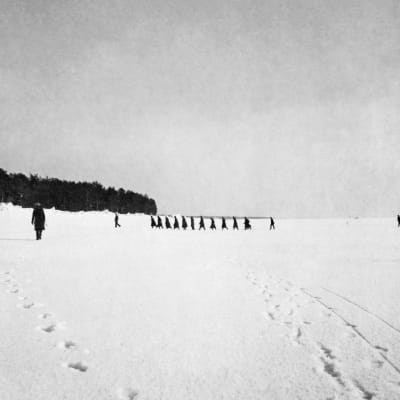 Svartvitt foto från 1921. Svarta skepnader på havsisen nära stranden. De första flyktingarna kommer.