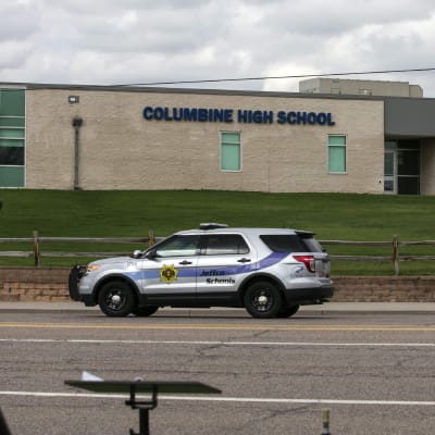 En bild på Columbine High Schools sandfärgade byggnad. På vägen framför byggnaden kör en polisbil förbi.